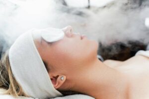 Blonde woman with headband having a facial steaming treatment, eyes covered with cotton pads, side view.