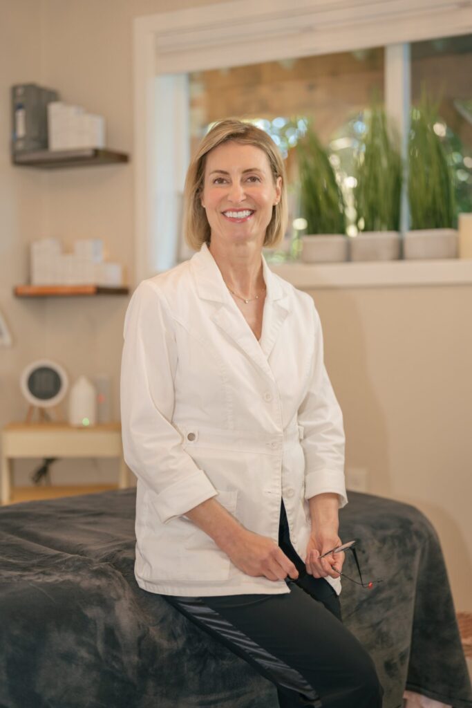 Smiling woman with short blonde hair wearing a white lab coat and holding glasses in a treatment room.