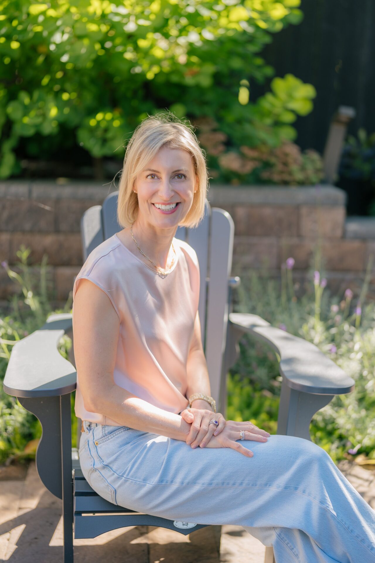 Nicole with a pink top sitting outside smiling in a gray Adirondack chair. Nicole provides skincare therapy Edmonds WA