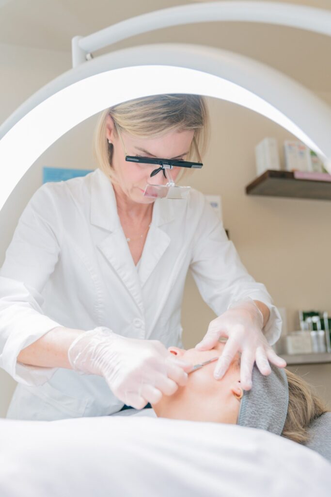Skincare Aesthetician wearing magnifying glasses performing a facial treatment on a client lying down.