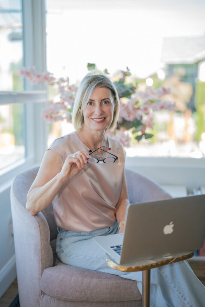 Woman with blonde hair smiles, holding glasses, with a laptop in front of her in a bright, airy room.