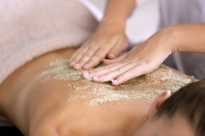 A woman receives a salt scrub on her back at a spa; hands apply the scrub. This is part of our enhancements.