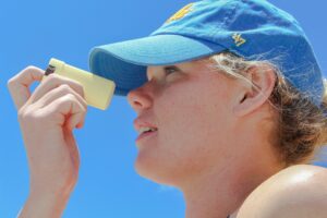 Woman wearing blue cap and applying sunblock to her face on a sunny day, with blue sky background.