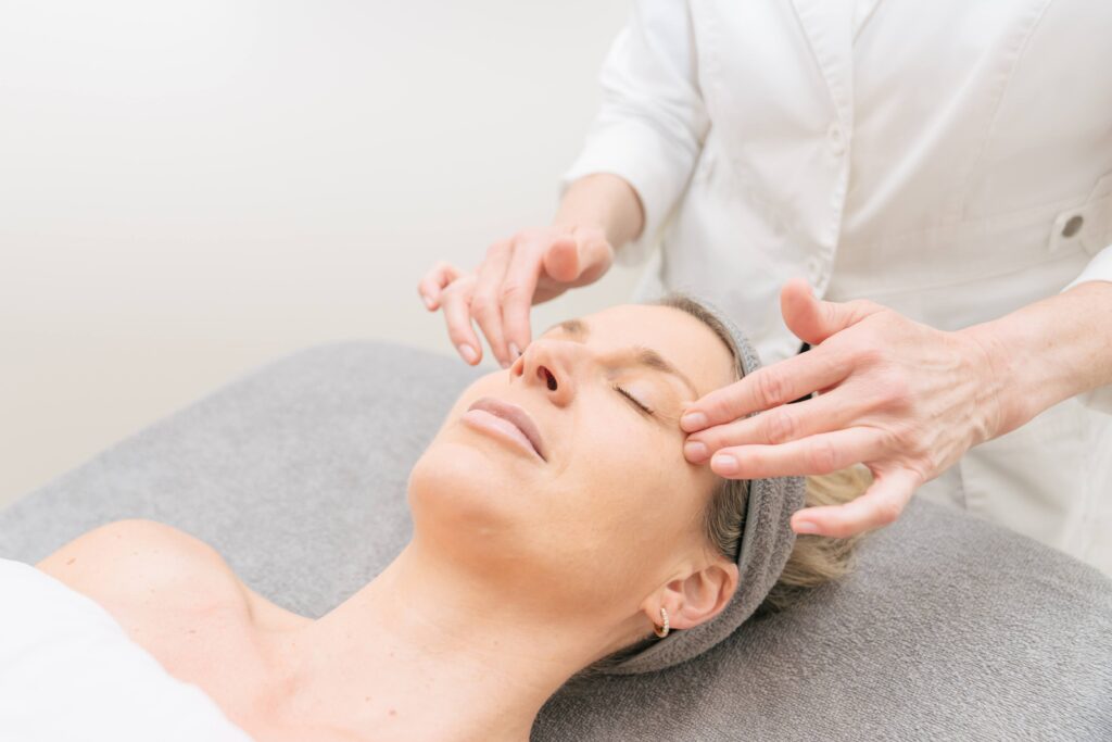 A woman receiving a facial massage while lying on a treatment table, eyes closed, wearing a gray headband.